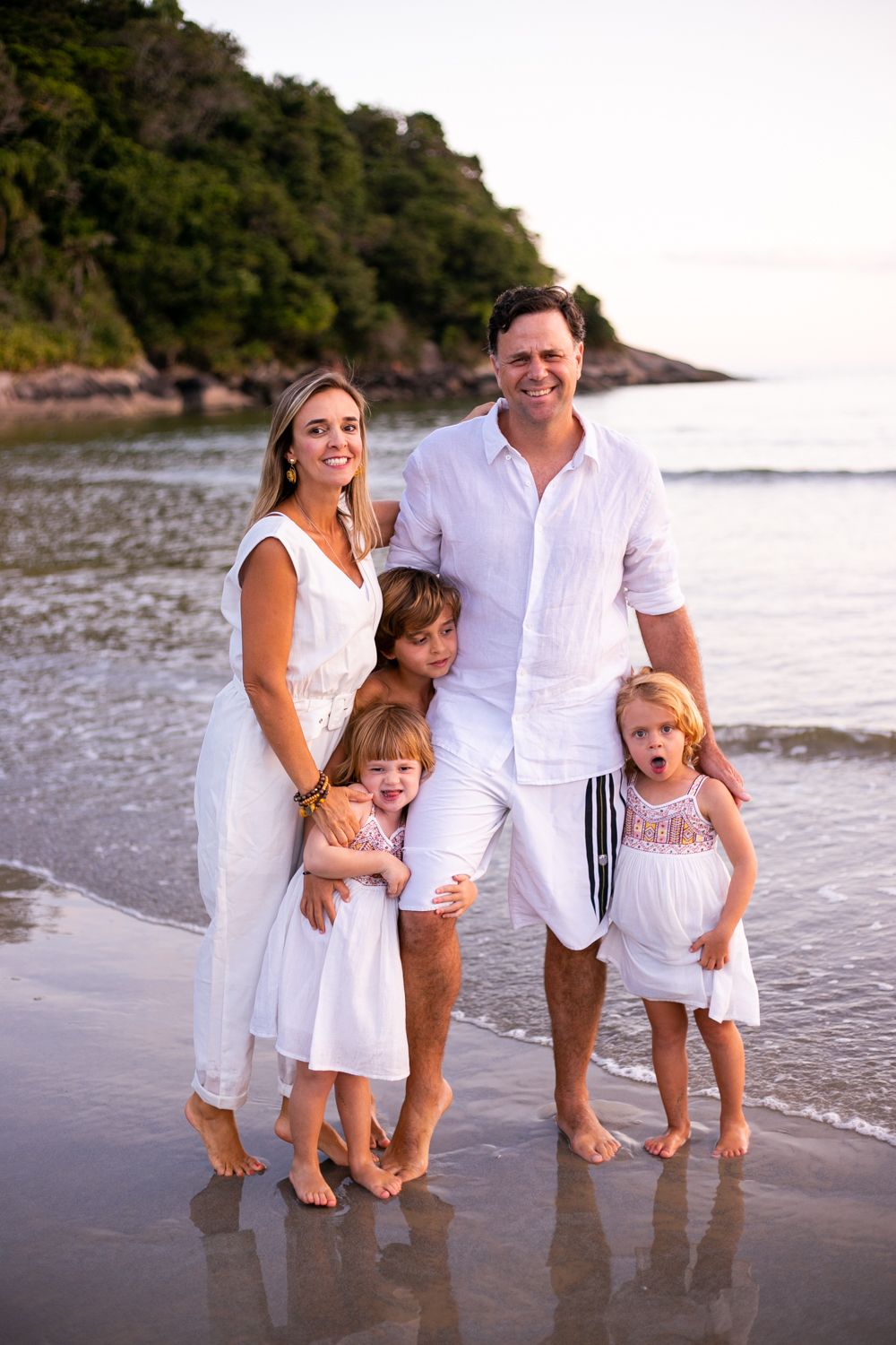 Family wearing white outfits poses together on a tropical beach at sunset with cliffs in the background.