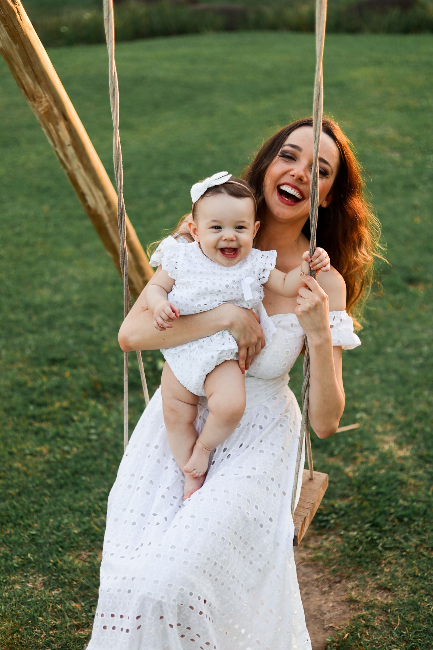 A joyful moment of mother and baby in matching white dresses playing on a wooden swing in a green garden.