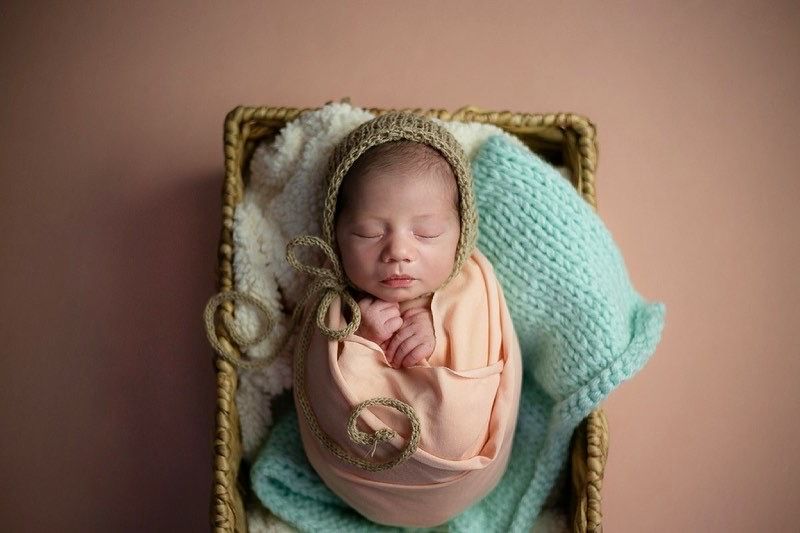 A sleeping newborn swaddled in peach fabric rests in a woven basket with a mint green knit blanket.