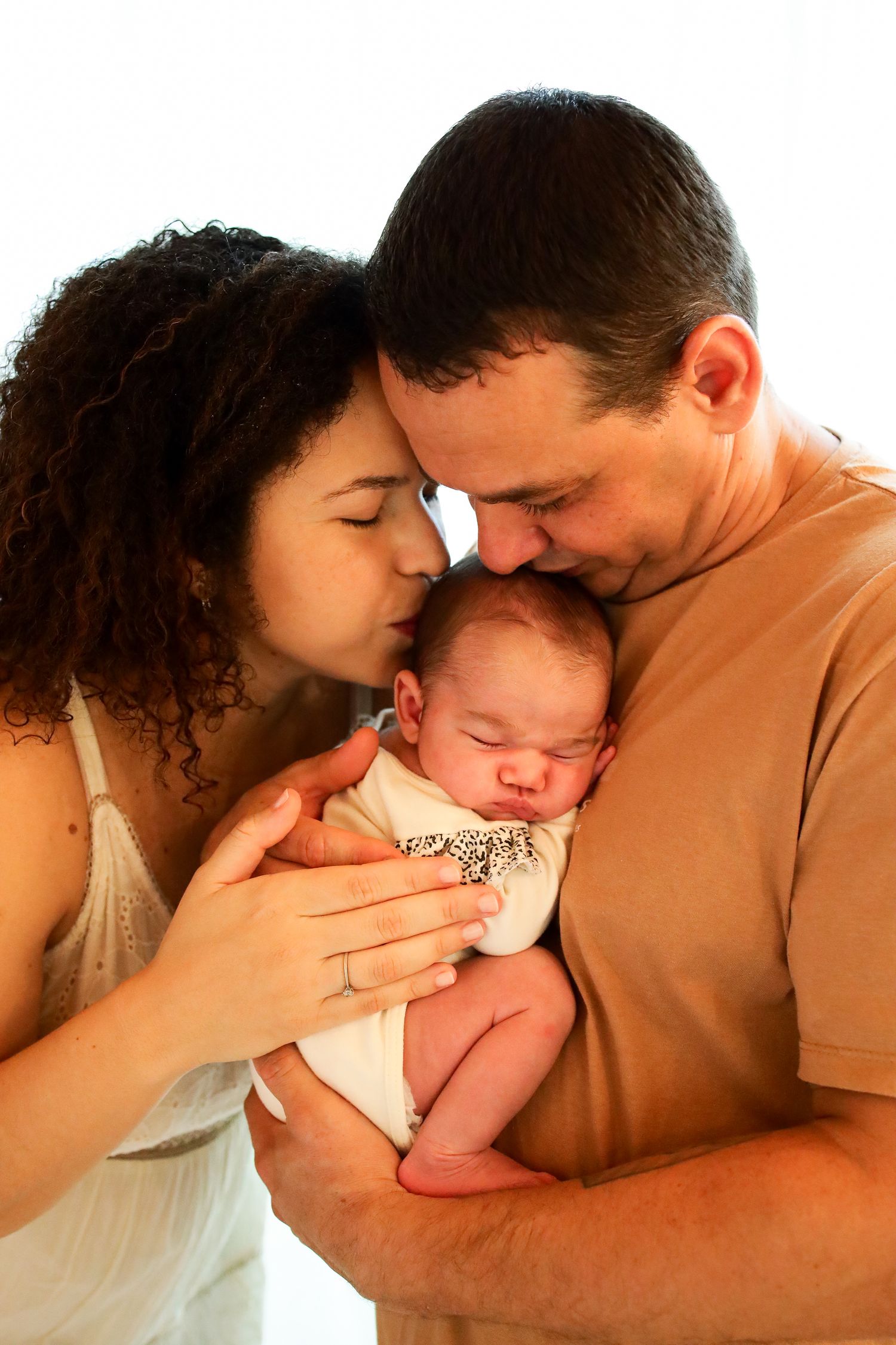 A loving couple tenderly embraces their sleeping newborn baby in warm lighting.