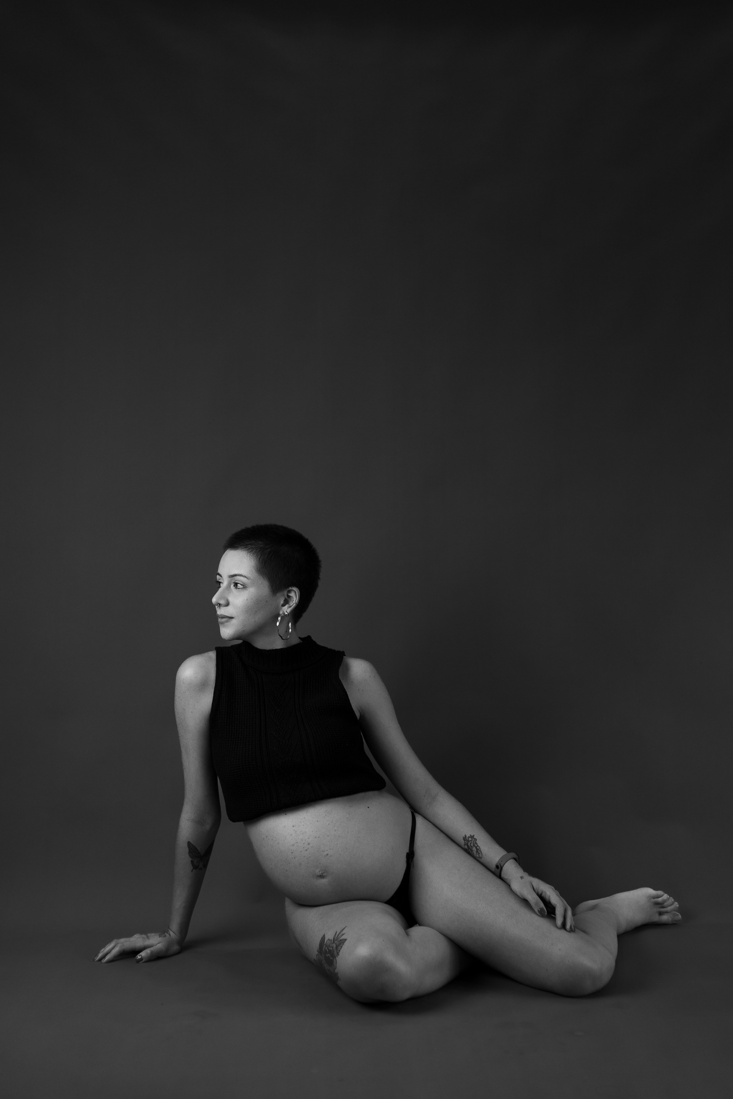 Artistic black and white maternity portrait of an expectant mother in a crop top seated on the floor.
