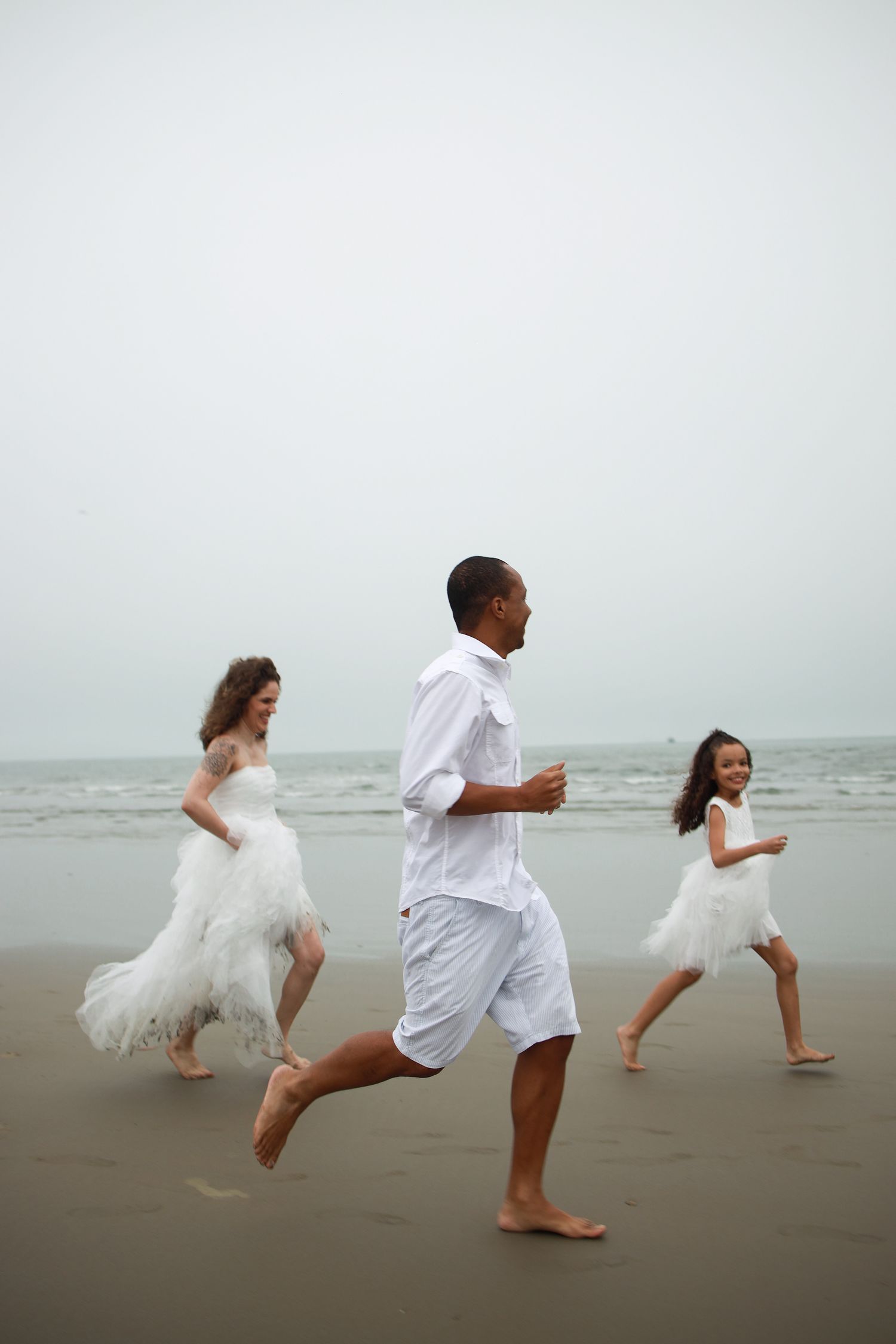 Three people wearing white running barefoot together on a misty beach along the shoreline.