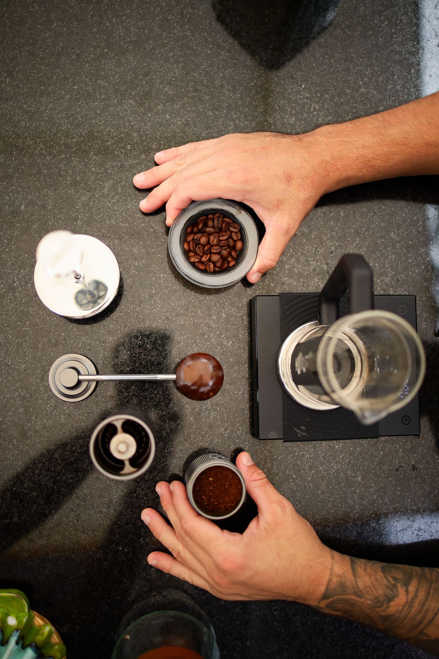 Overhead view of hands preparing coffee with beans, grinder and brewing equipment on dark surface.