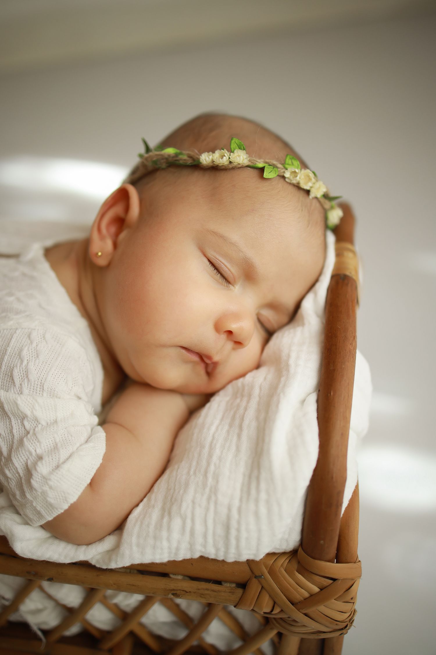 Sleeping baby wearing white dress and floral crown rests peacefully in woven basket with soft white bedding.