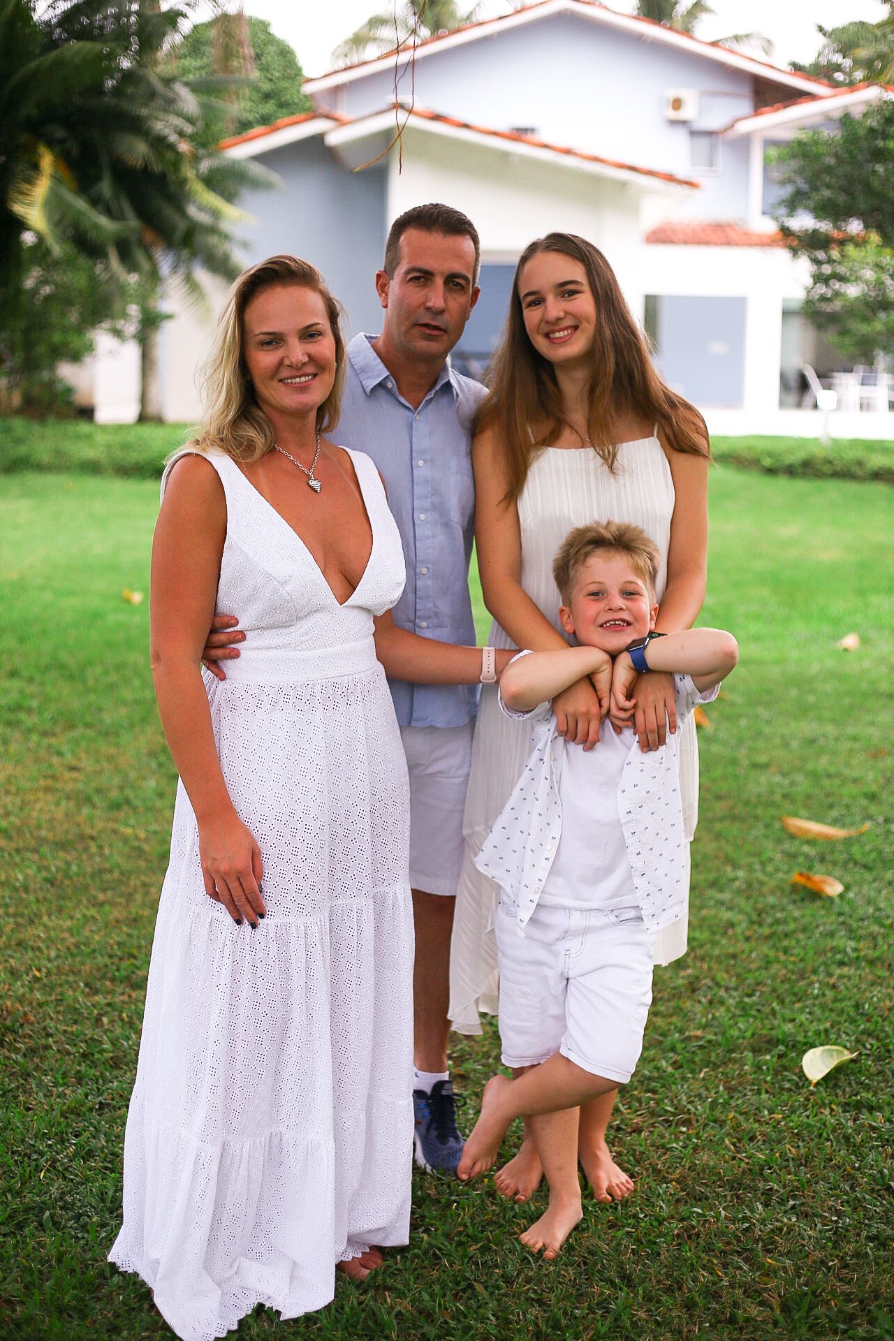 A family wearing white outfits poses together on a sunny lawn in front of a house.