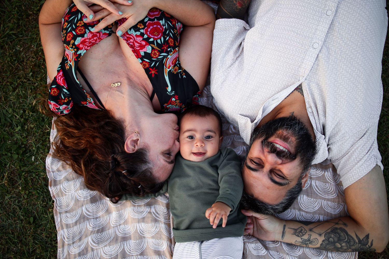 A baby in a gray outfit lays on a blanket in the grass between two adults who are laying down.