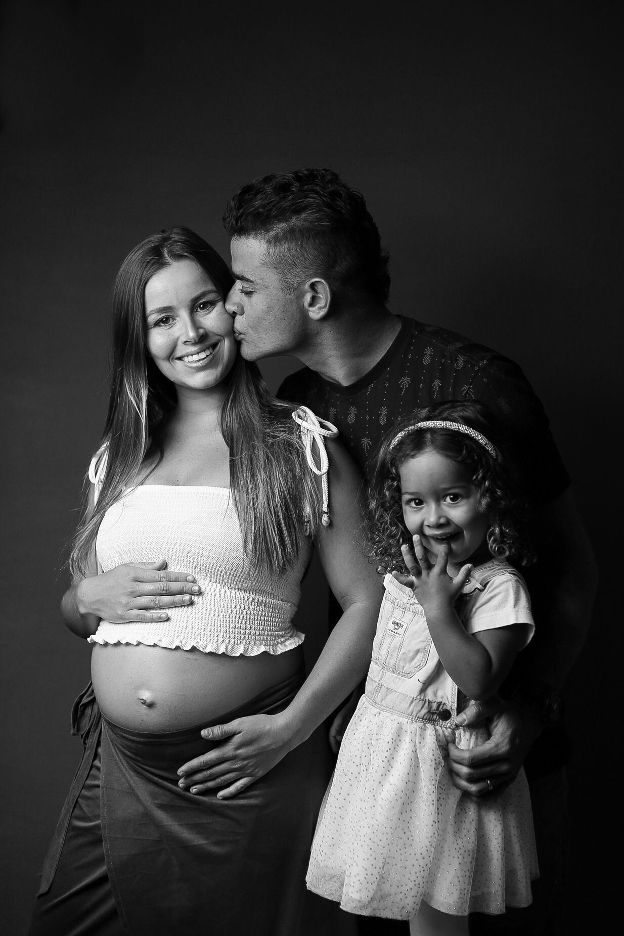 Black and white portrait of a pregnant woman in white top with a little girl in a white dress standing together against dark backdrop.