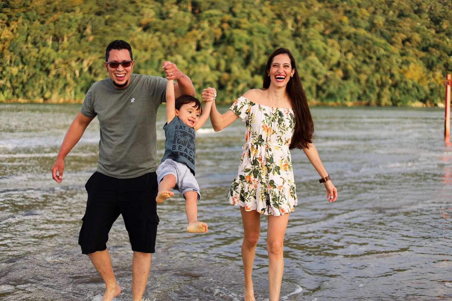 A family enjoys a playful moment together while walking in shallow water on a sunny day by a forested shoreline.