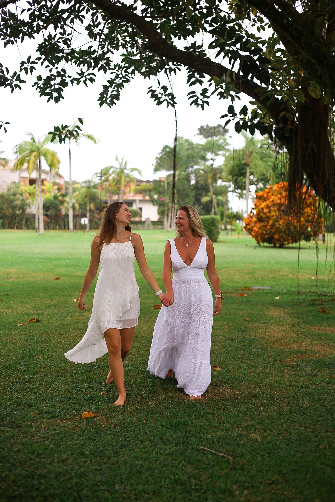 Two people in white dresses walk hand in hand on a lush green lawn under a tree with palm trees in the background.