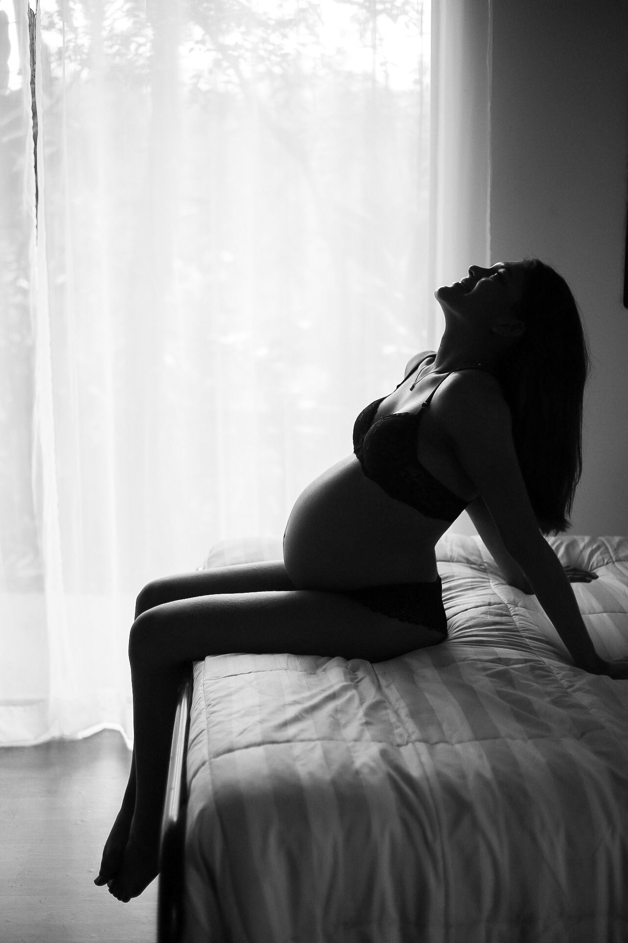 Silhouette of an expectant mother in black lingerie reclining on a bed near a window in dramatic black and white.