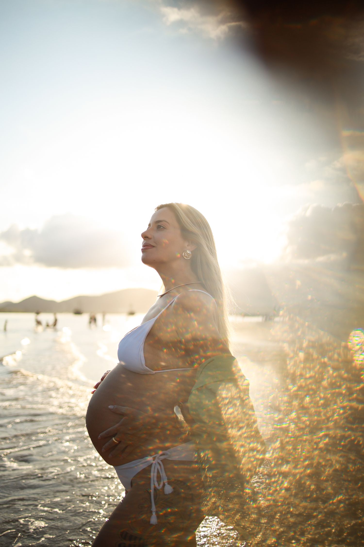 Silhouette of pregnant woman standing on beach during golden hour sunset with light flares.