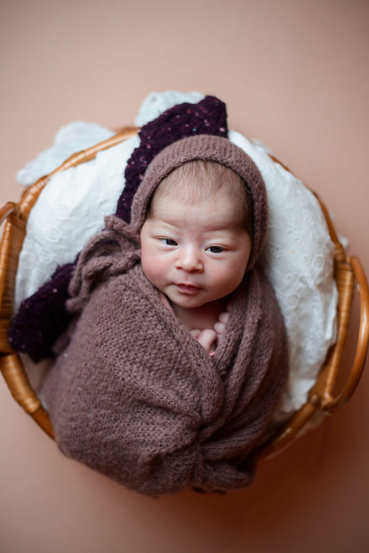 A tiny baby wrapped in brown knit blanket rests in a woven basket with white padding.