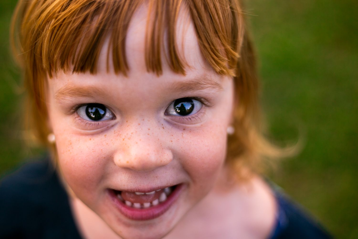 A close-up portrait of a smiling child with bright blue eyes and reddish hair against a blurred green background.