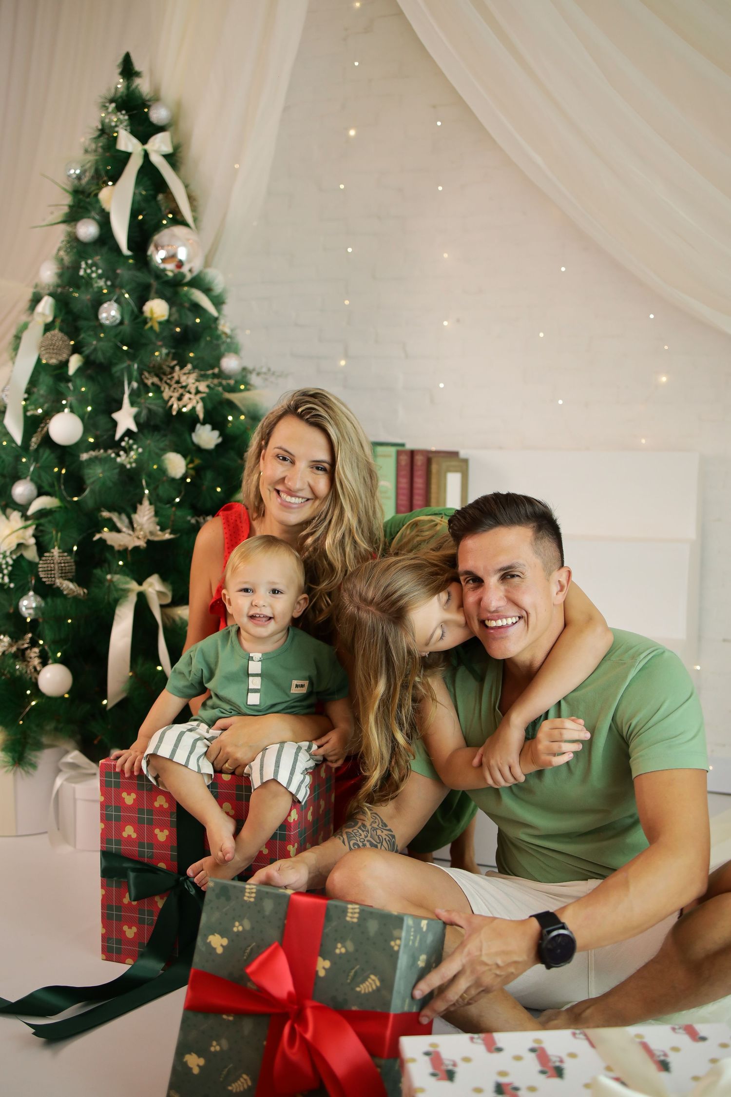 Family poses together in matching green outfits next to decorated Christmas trees and presents in a festive holiday setting.
