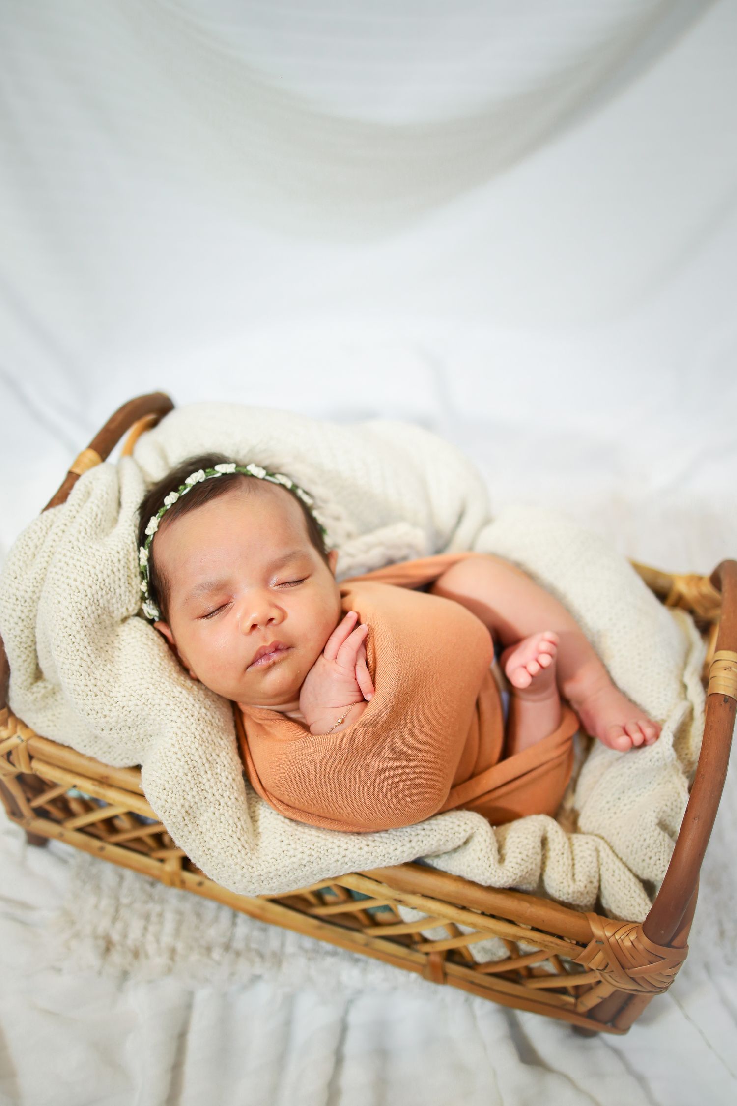 Sleeping newborn in orange wrap rests peacefully in woven basket lined with cream colored blankets.