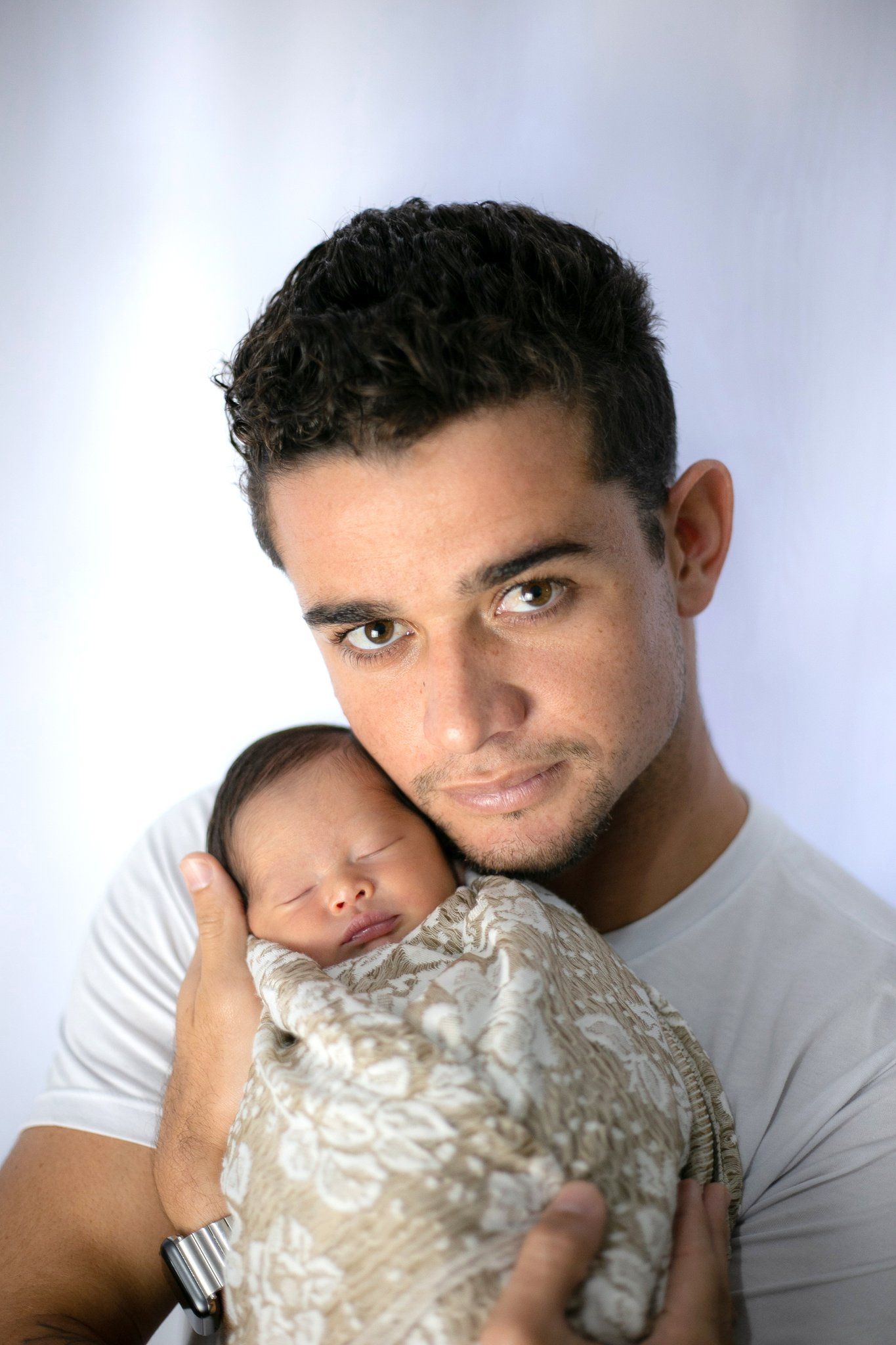 A young man in a white t-shirt tenderly holds a sleeping newborn baby wrapped in a beige blanket.