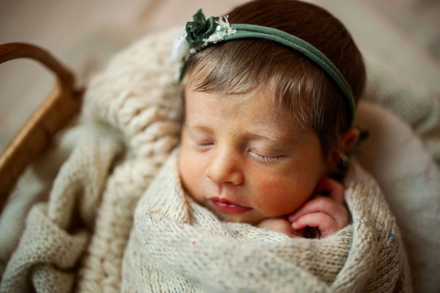 Sleeping newborn wrapped in cream knit blanket wearing mint green headband nestles peacefully during photo session.