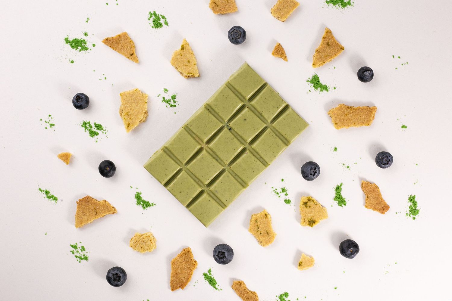Green matcha chocolate bar surrounded by scattered blueberries, cookie crumbles and fresh herbs on white background.