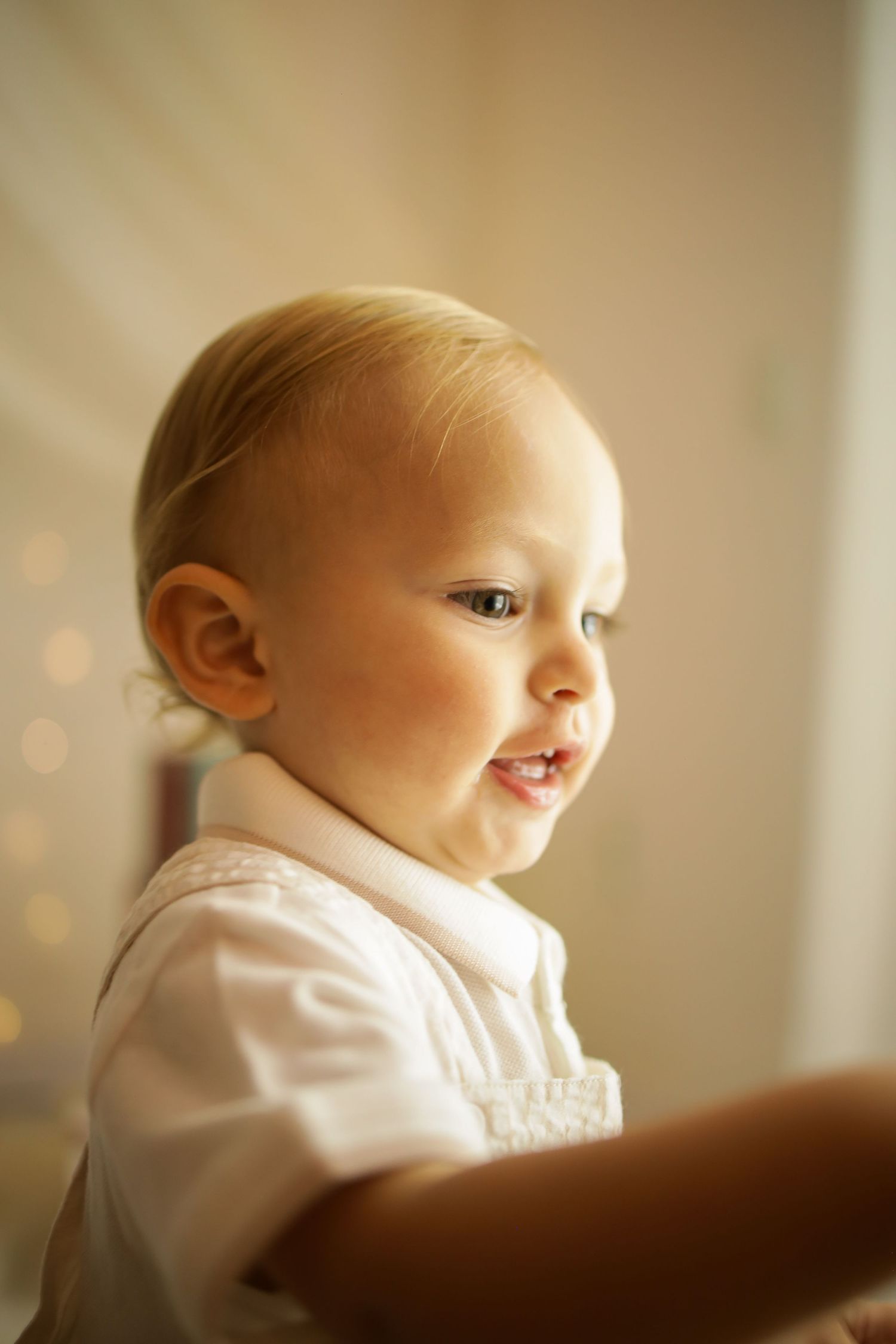 A baby in a white shirt gazes softly to the side in warm natural light creating a gentle portrait.