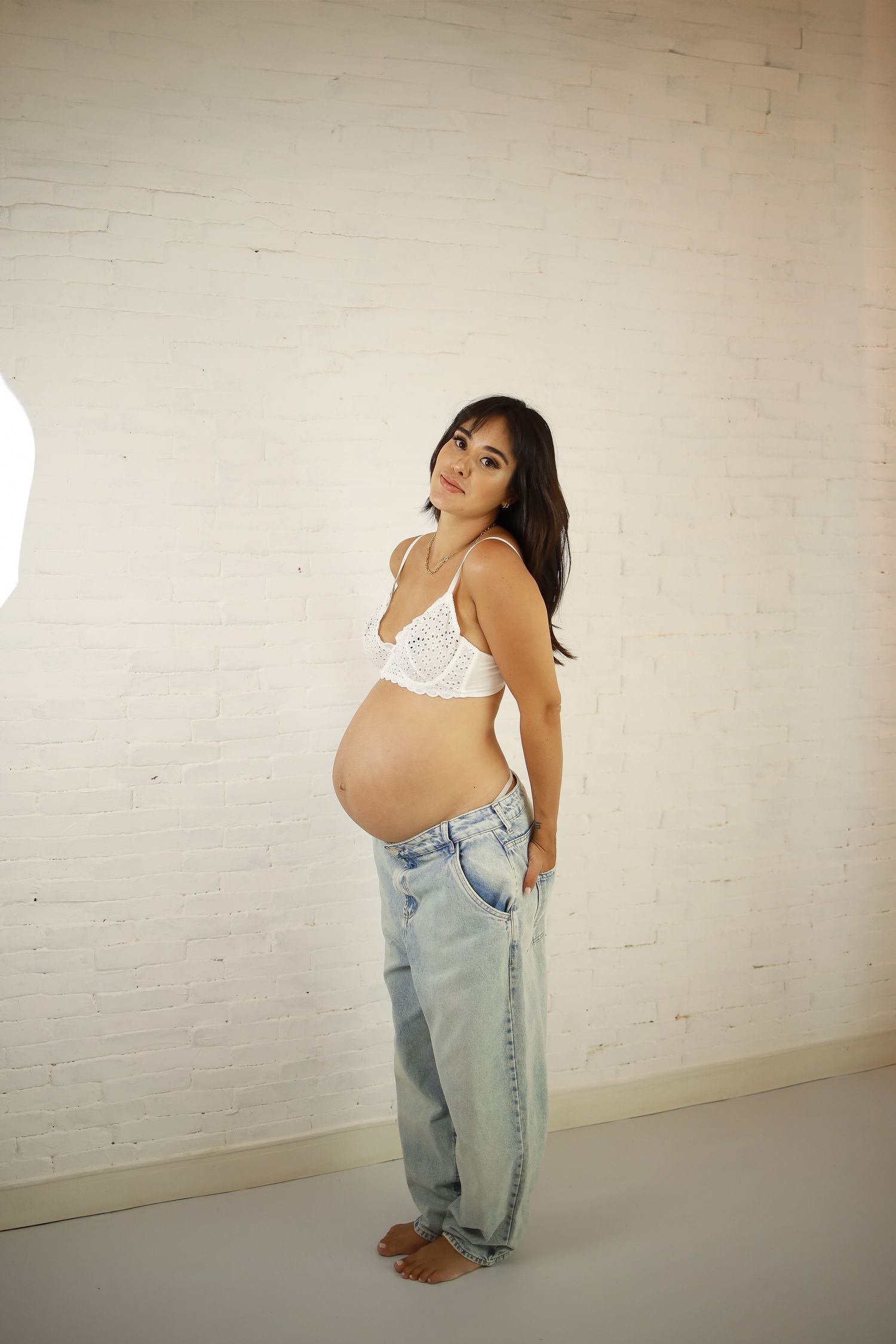 Expecting mother in loose jeans and white bra top poses against white wall for maternity photo.