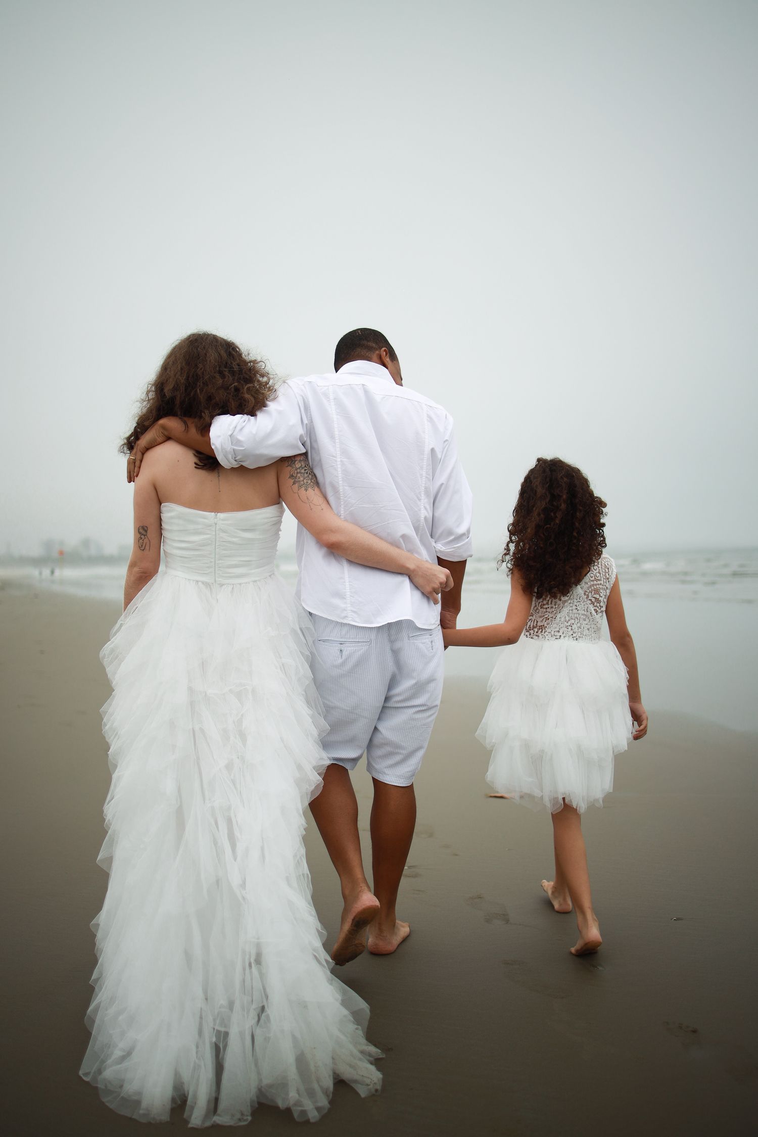 Three people walking together barefoot on a beach, with two wearing flowing white dresses and one in a white shirt and shorts.
