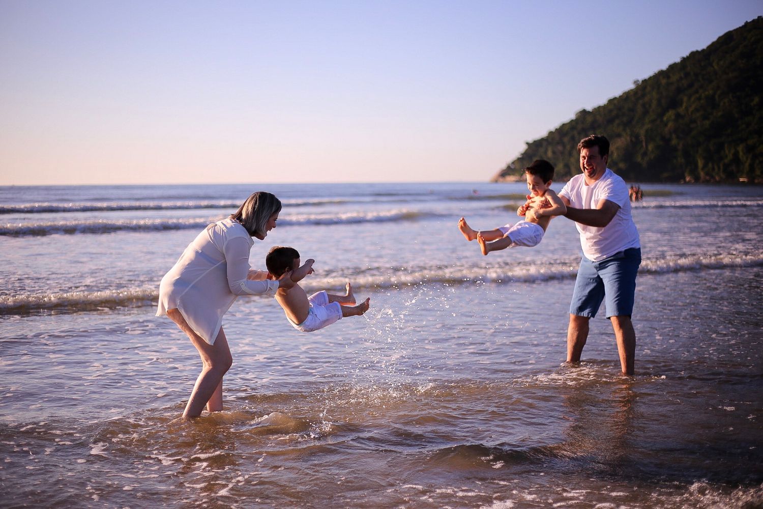 People playing and splashing in the ocean waves at sunset along a scenic beach with mountains in the background.