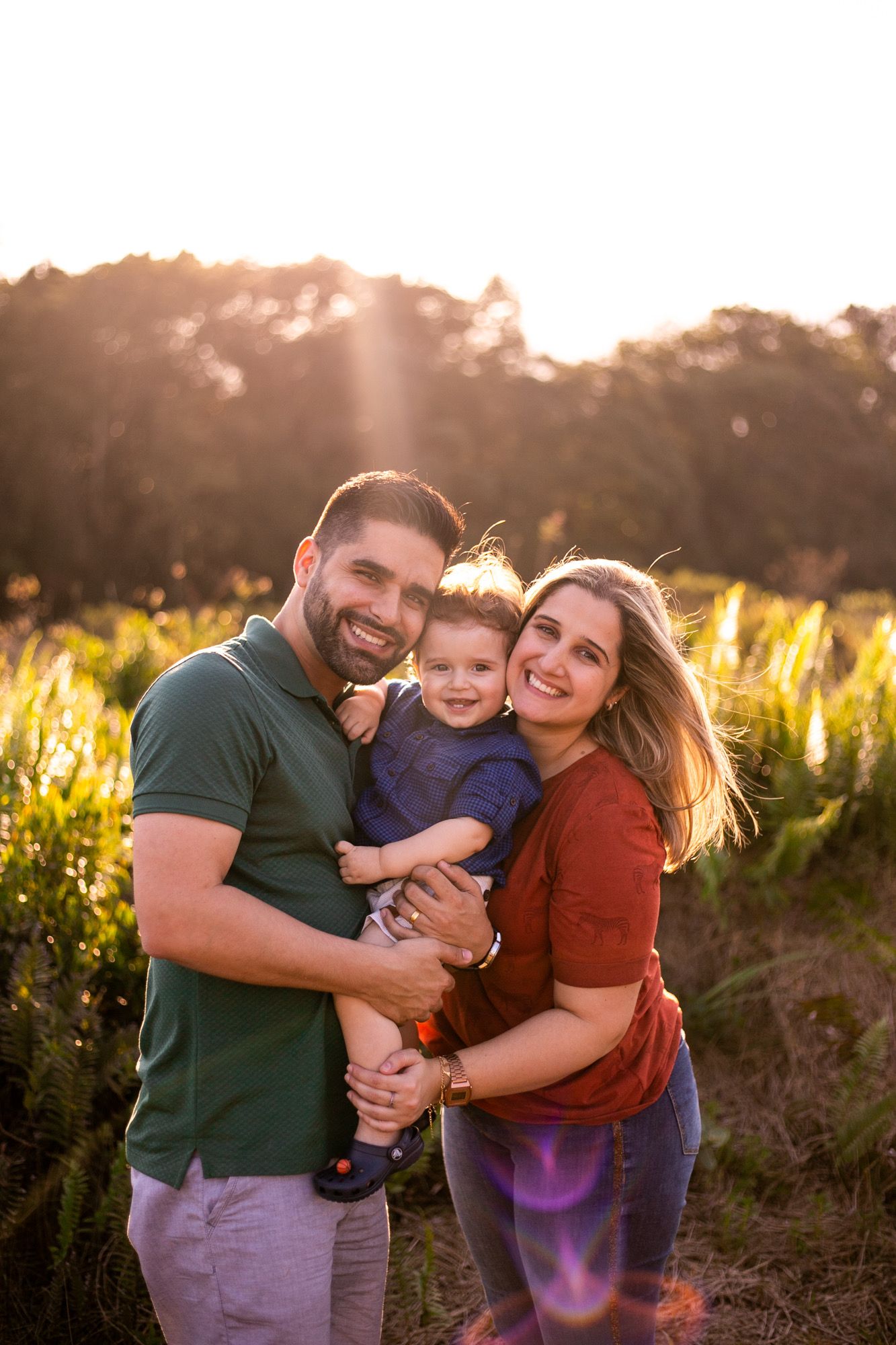 Family stands together in a golden sunlit field during sunset, embracing and smiling at the camera.