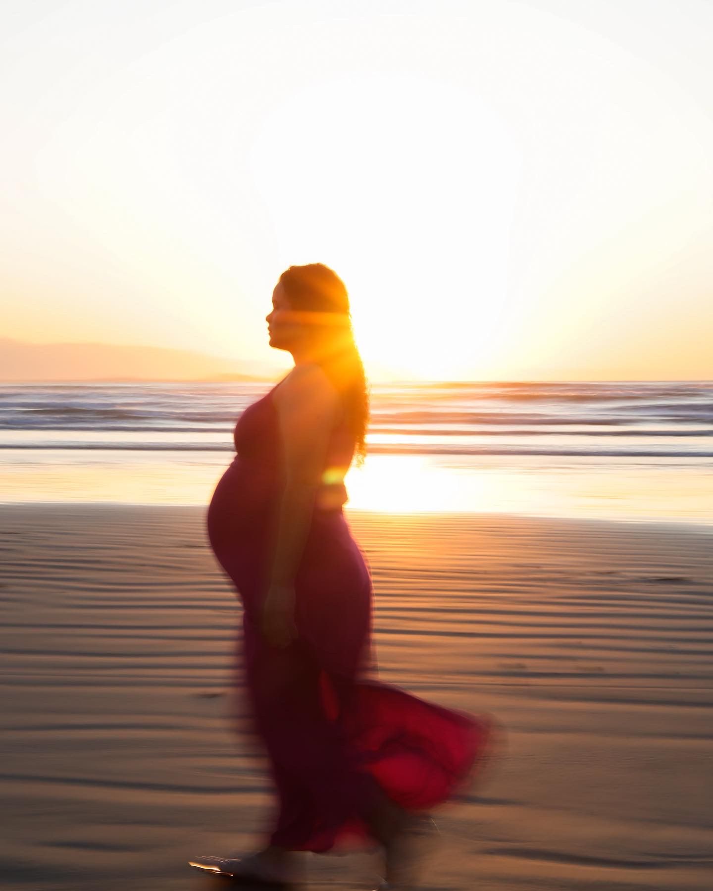 A silhouette of a pregnant figure in a flowing dress standing on a beach at sunset with golden light streaming behind.