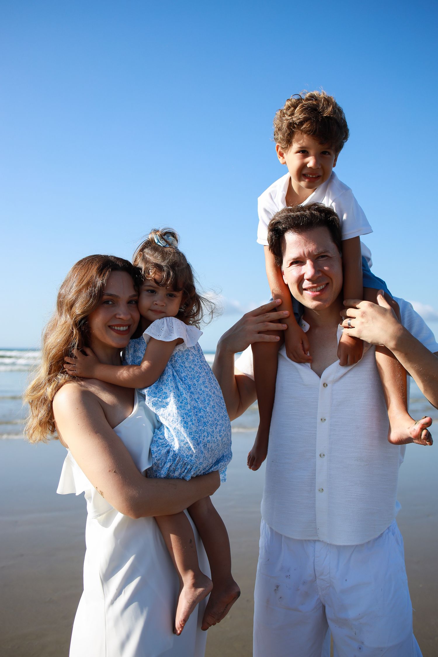 A family wearing white outfits poses together for candid photos on a sunny beach with blue skies in the background.
