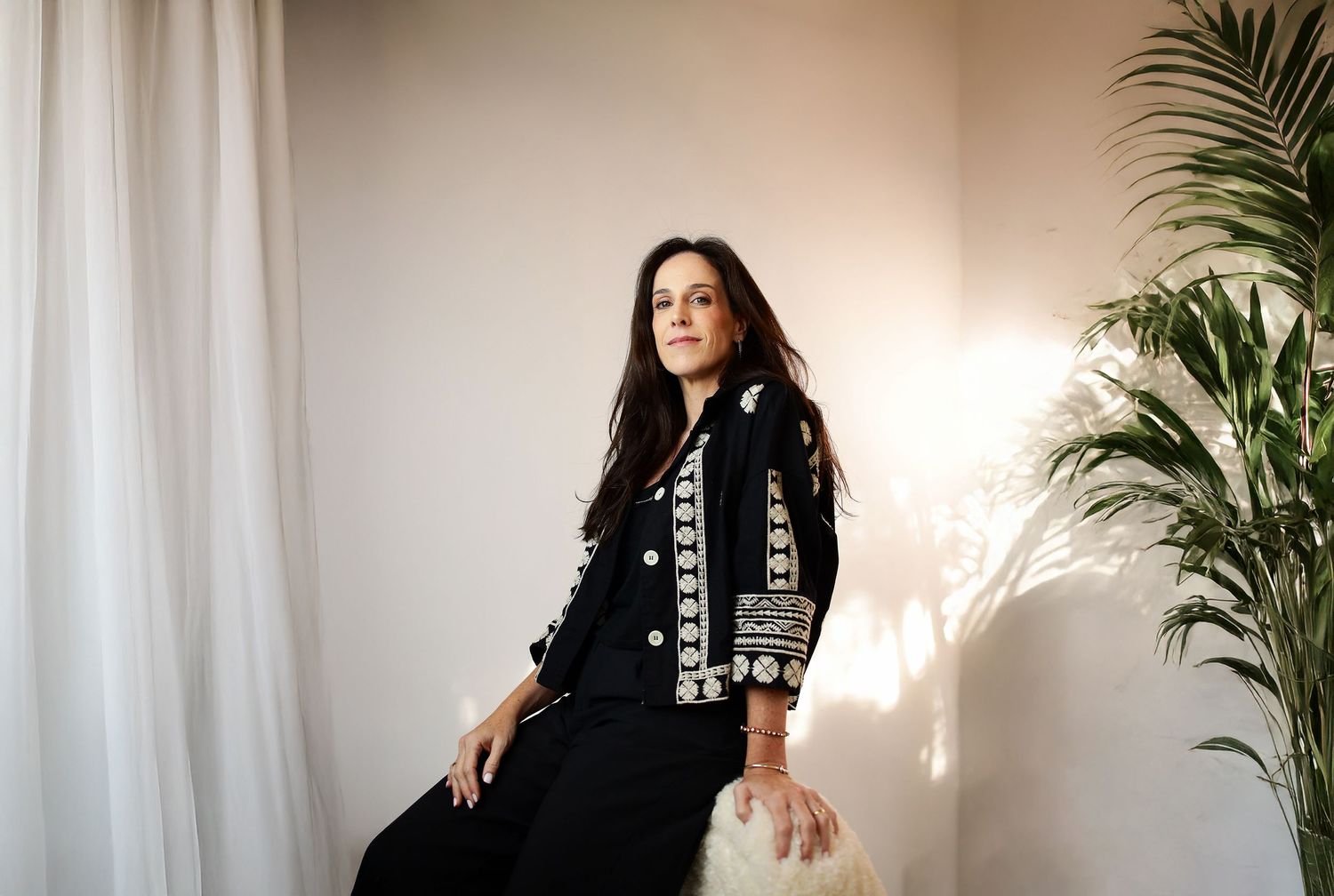 A person in a black jacket with white embroidery sits near a white curtain and palm plant in soft natural lighting.