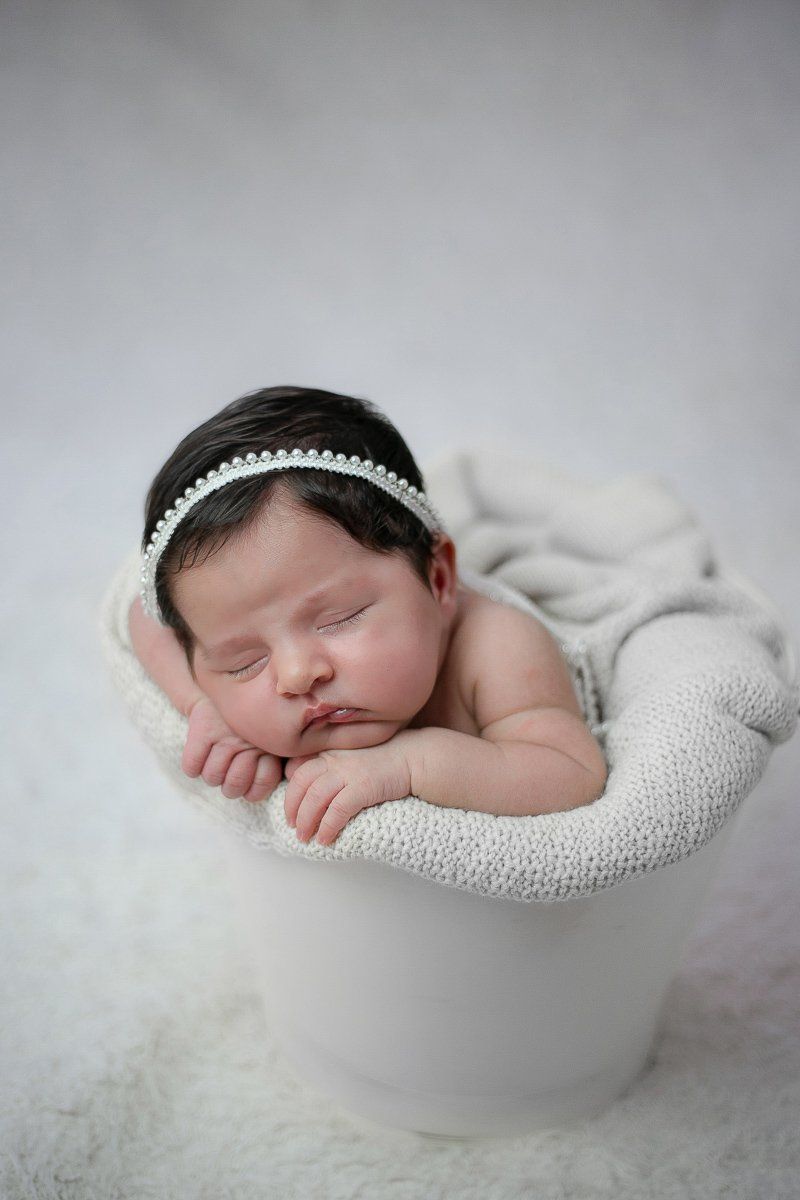Peaceful sleeping newborn wearing white headband rests on soft white fabric in white bowl against light background.