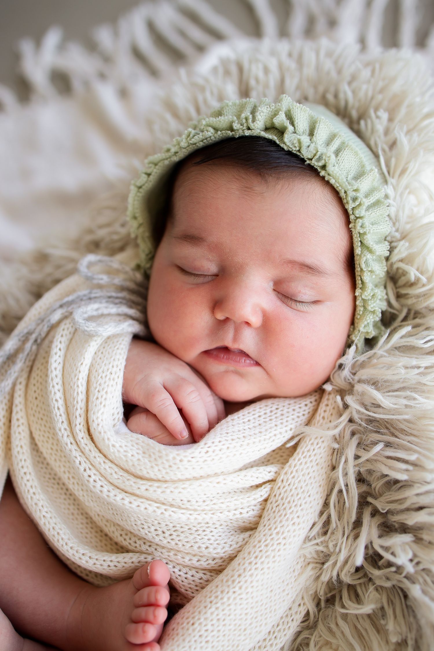 Sleeping infant wrapped in white knit blanket wearing mint green bonnet rests on fluffy cream textured backdrop.