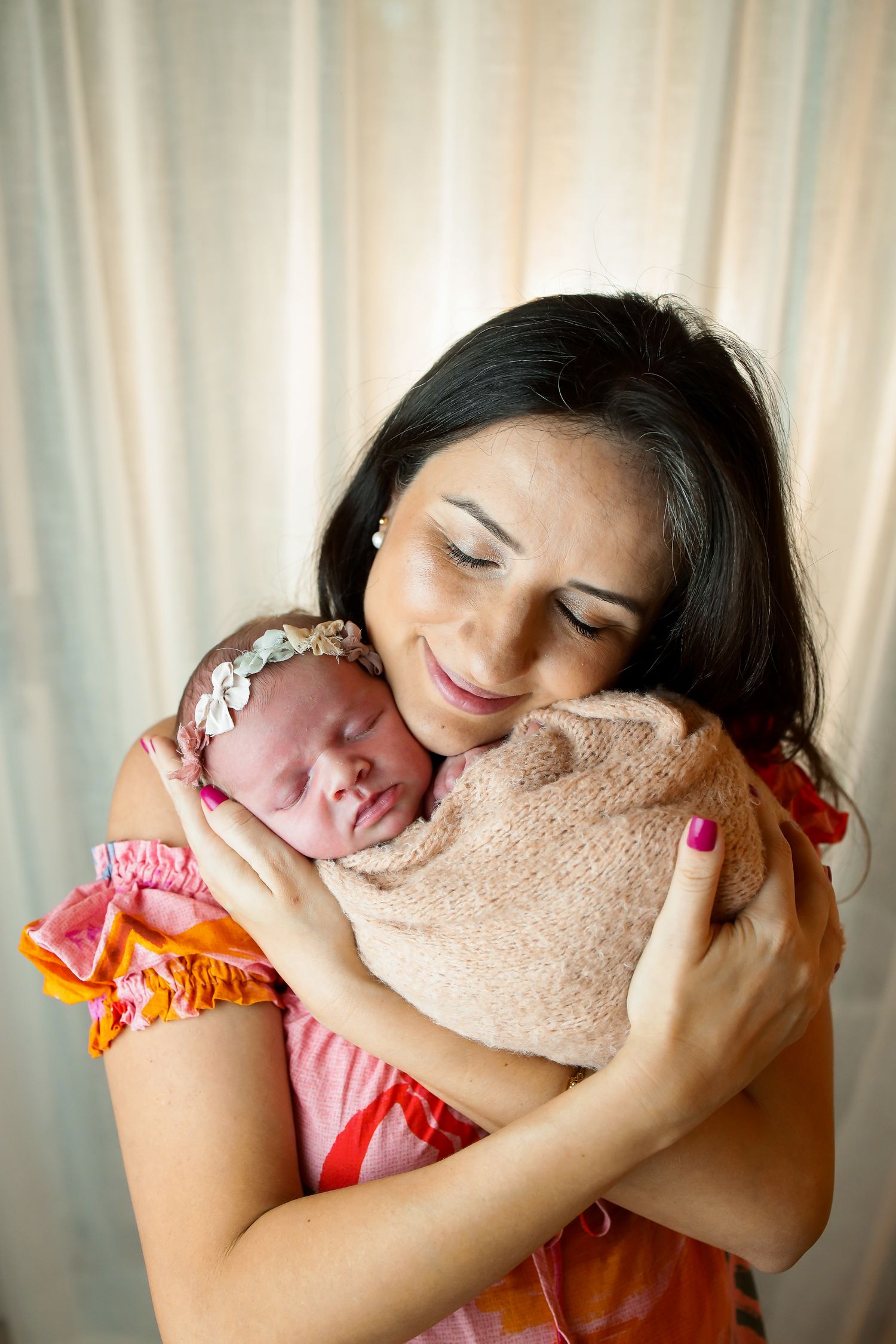 A tender moment as mother in coral dress cradles her sleeping newborn wrapped in a soft beige blanket.