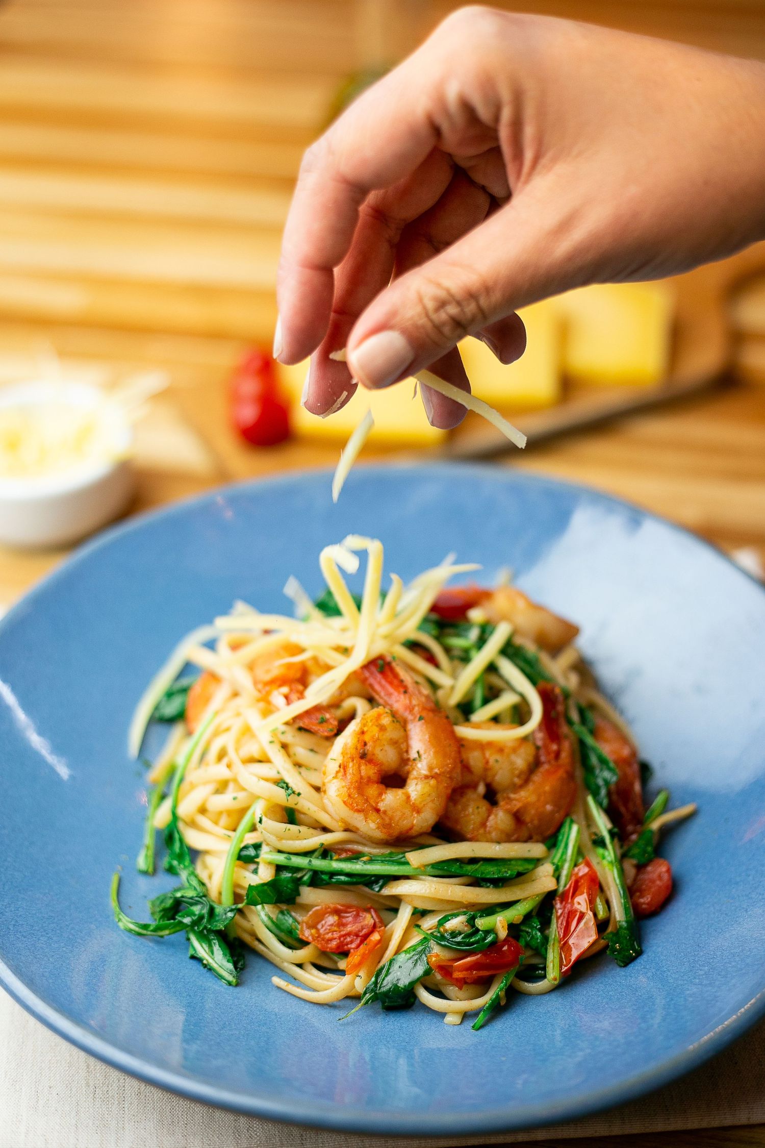 Hand grating parmesan cheese over a blue plate of spaghetti with shrimp, chili peppers and fresh herbs.