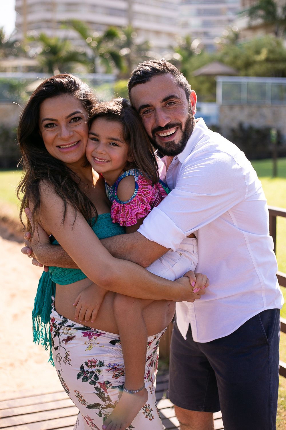 Family embraces together outdoors on sunny day with palm trees and buildings in background.