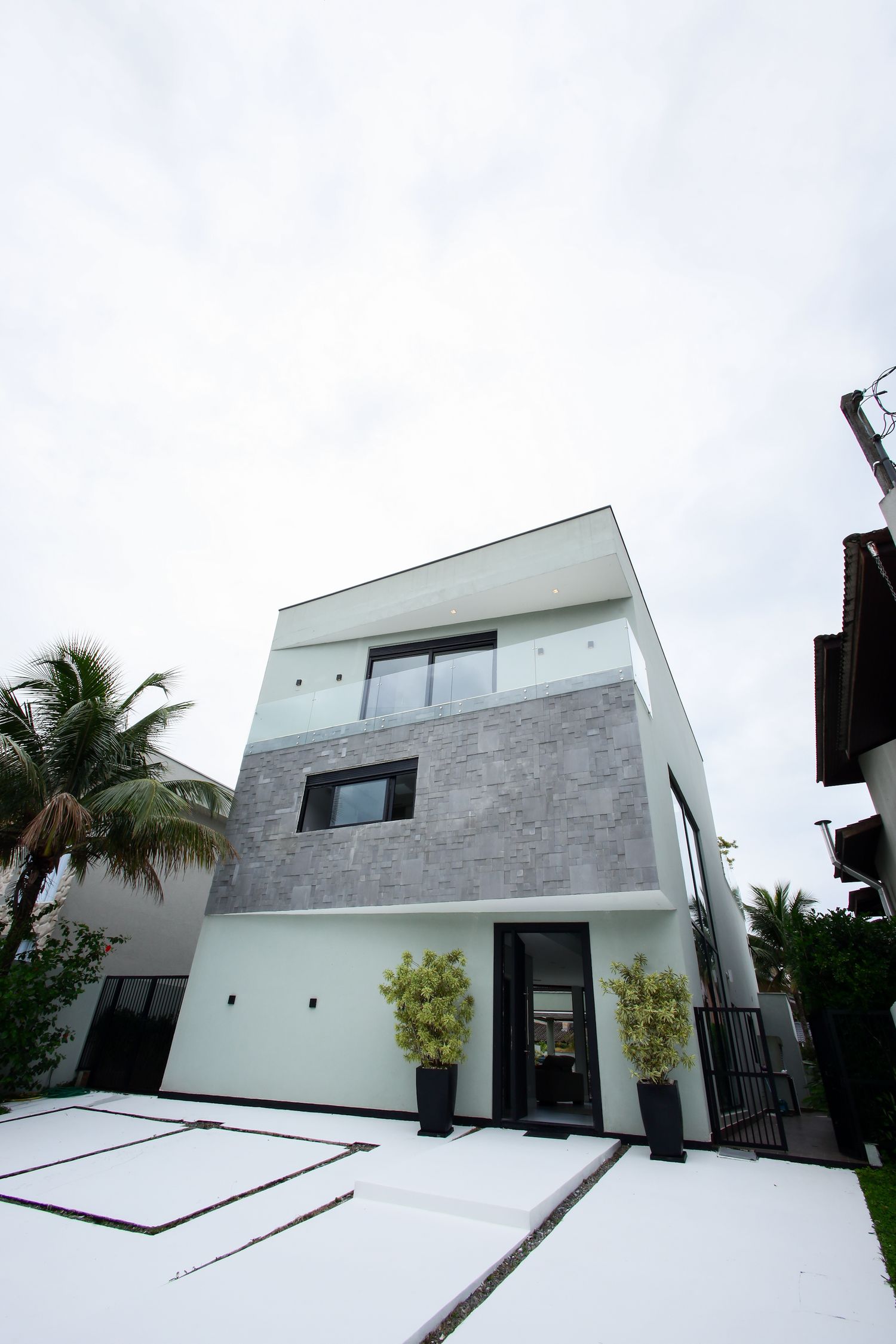 Modern three-story home with white and gray exterior, palm trees, and minimalist landscaping on a bright day.