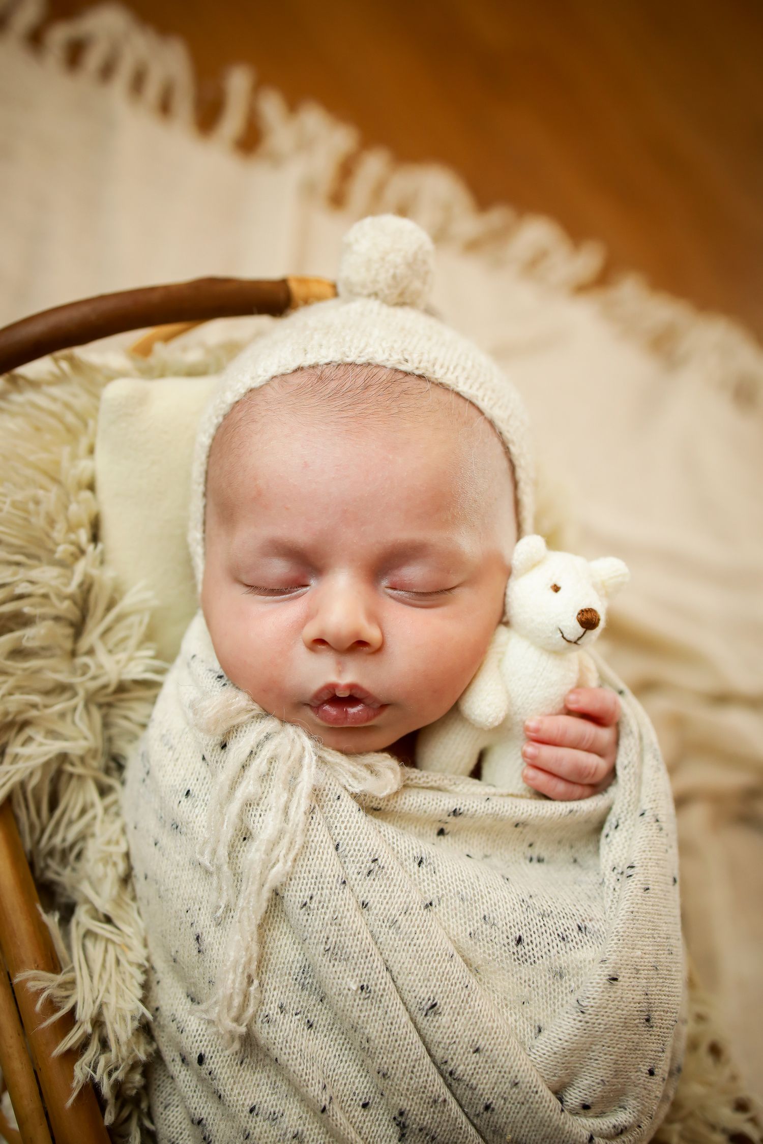 Sleeping baby wearing white knit sweater and hat holding small teddy bear while resting on soft blanket.