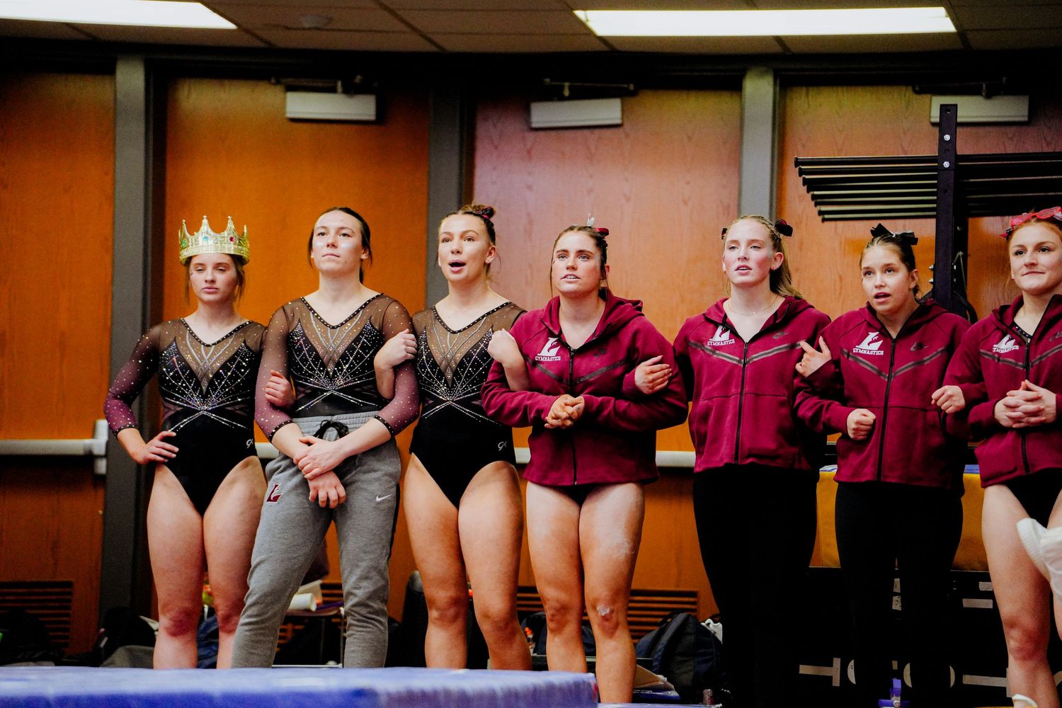 College gymnastics team members in black and burgundy leotards stand together watching a competition from sidelines.