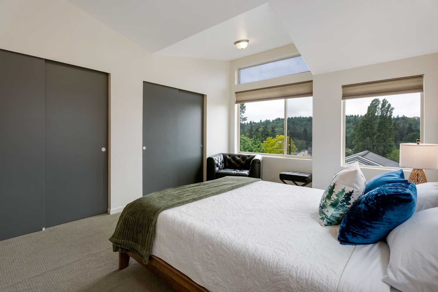 Modern bedroom with white king bed, teal accent pillows, gray closet doors, and large windows with tree views.