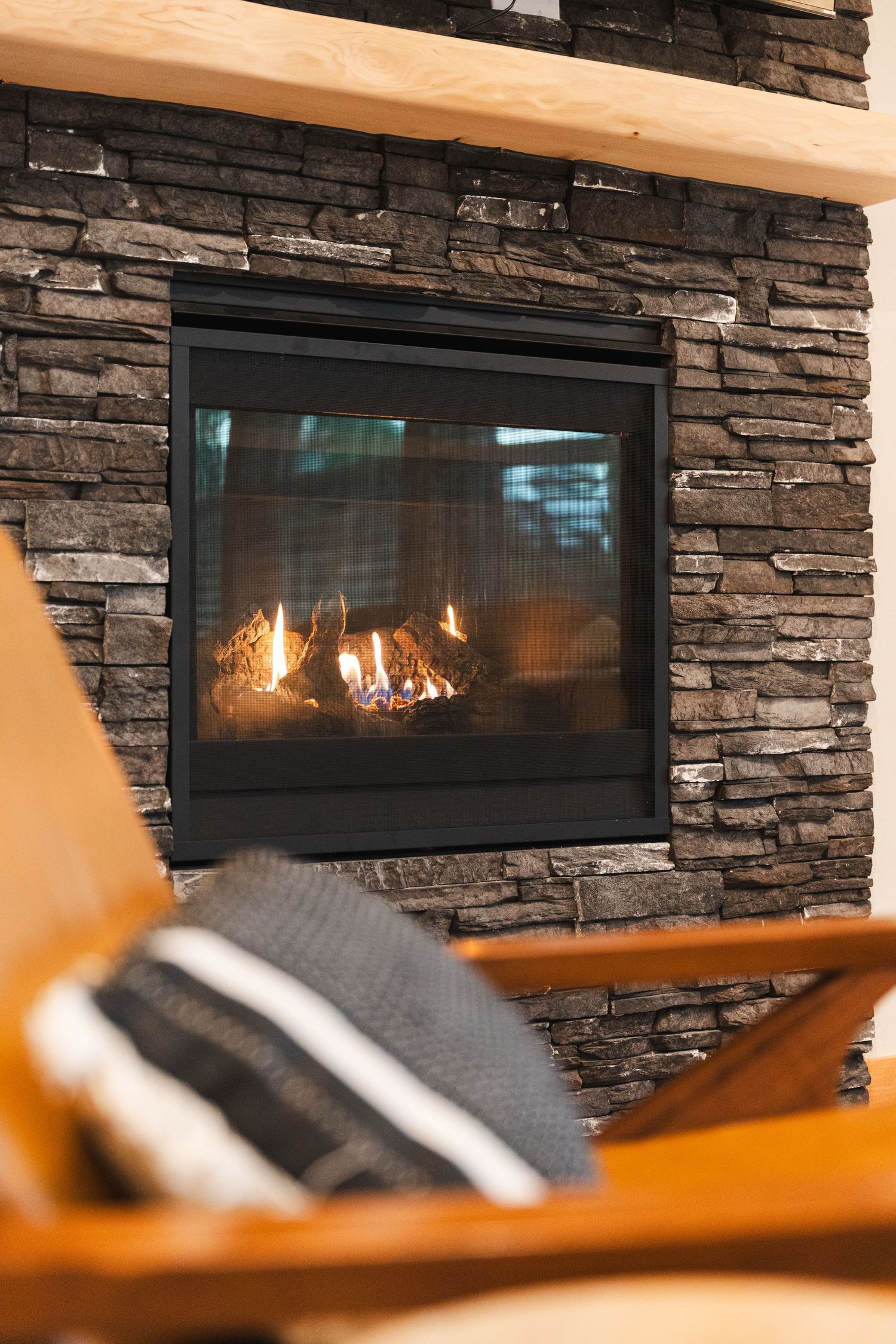 A cozy stone fireplace with burning flames, framed in black metal, with a wooden chair and striped pillow in the foreground.