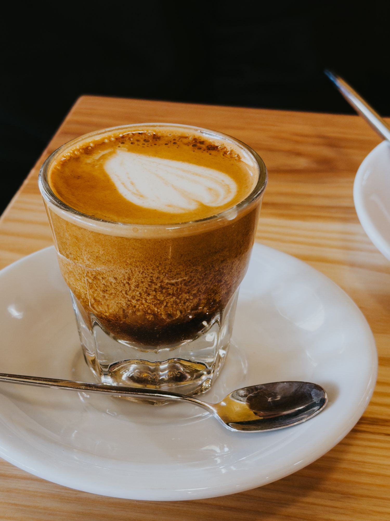 A small glass of layered espresso cortado with latte art sits on a white saucer with a spoon.