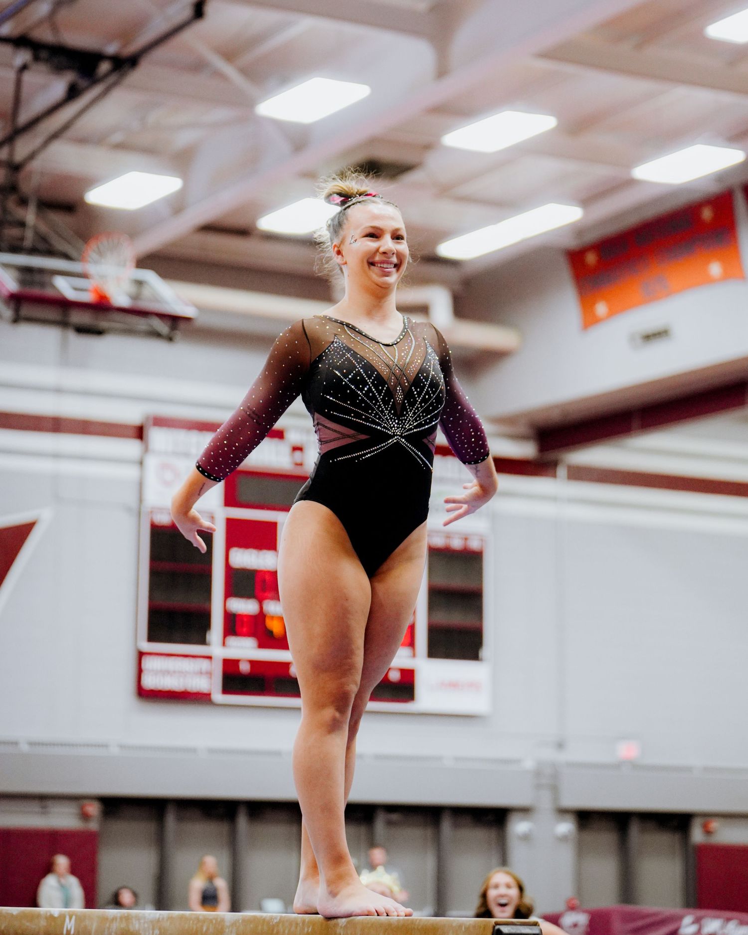 Gymnast performs on balance beam wearing black leotard with sparkly design in indoor gymnasium.