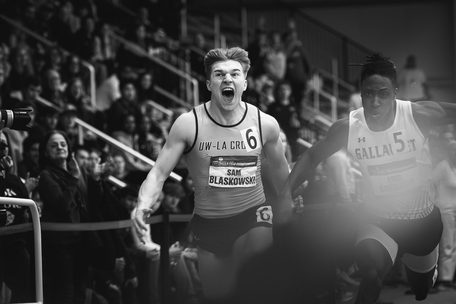 Black and white action shot of athletes competing in an indoor track event with spectators.