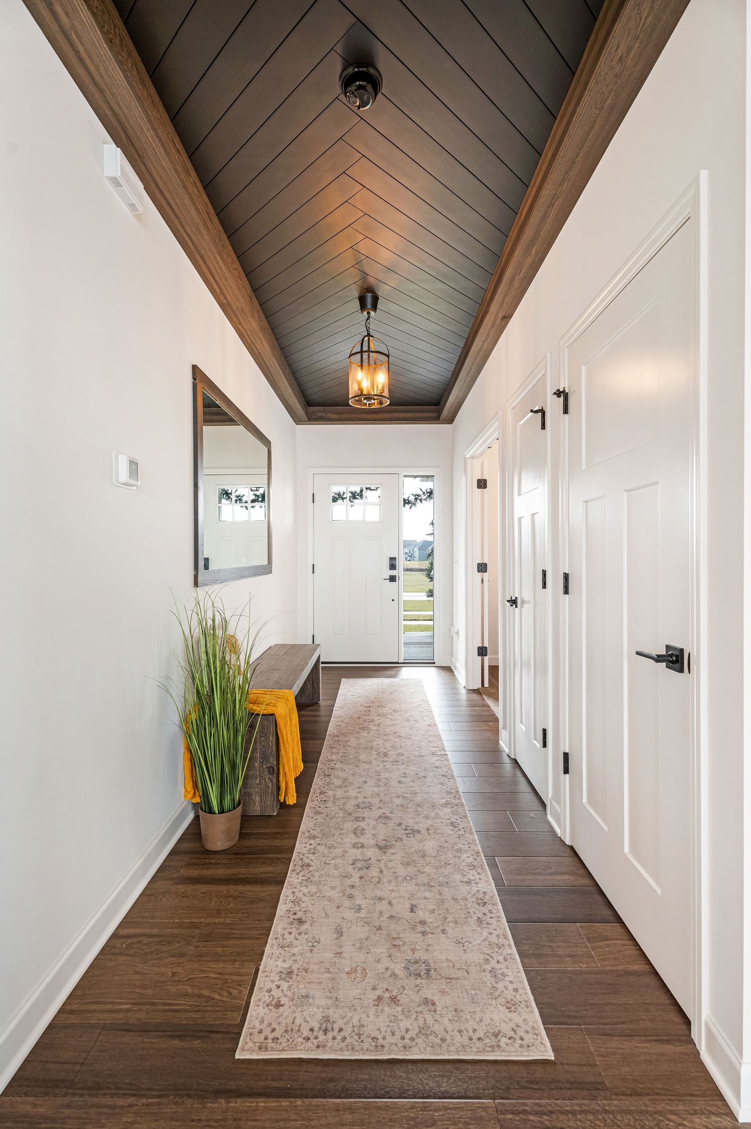 Elegant hallway with vaulted herringbone wood ceiling, pendant light, runner rug, bench, and white doors on dark hardwood floors.