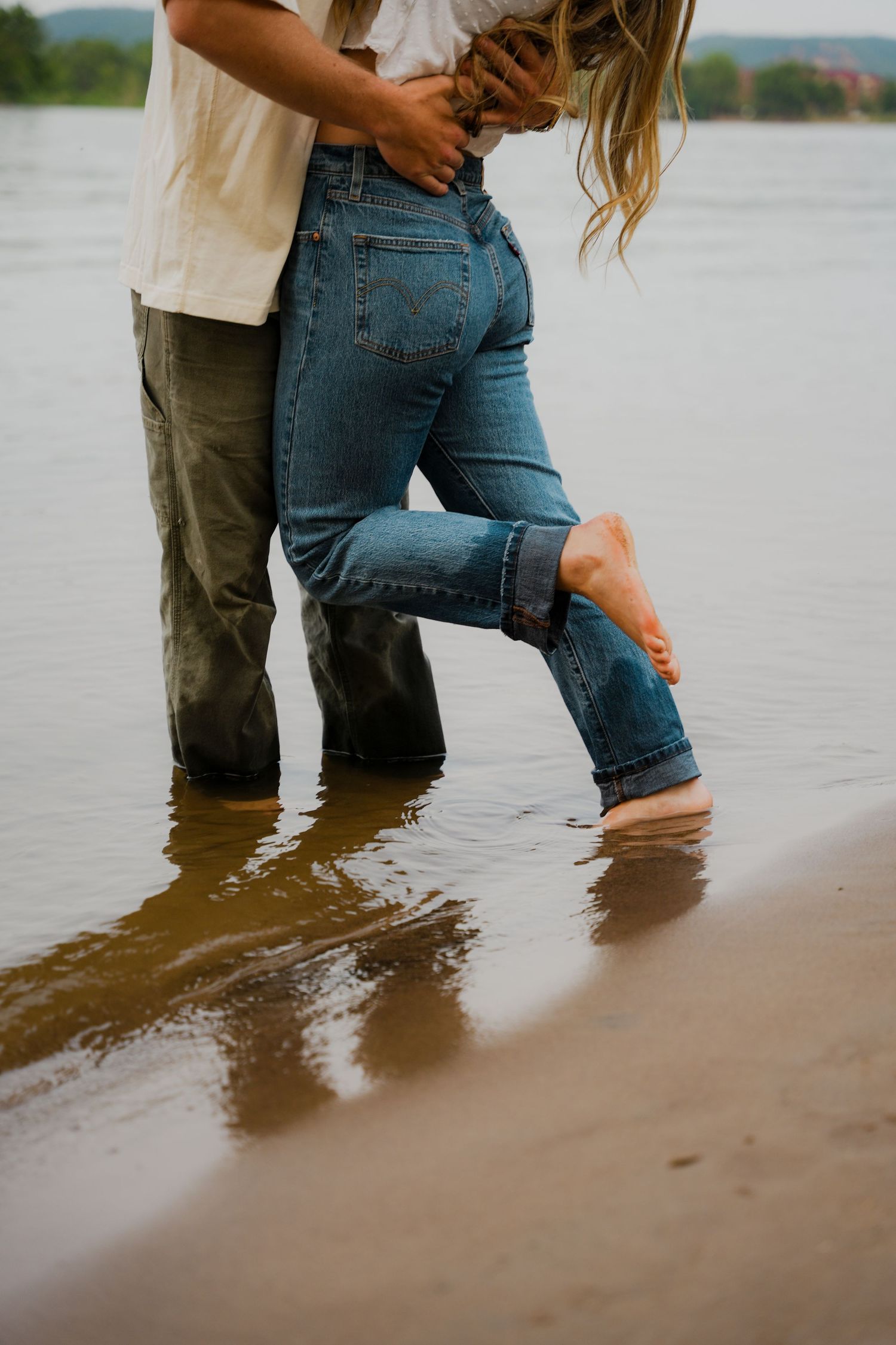 Two people standing in shallow water at the beach wearing jeans, with one person playfully lifting their leg.