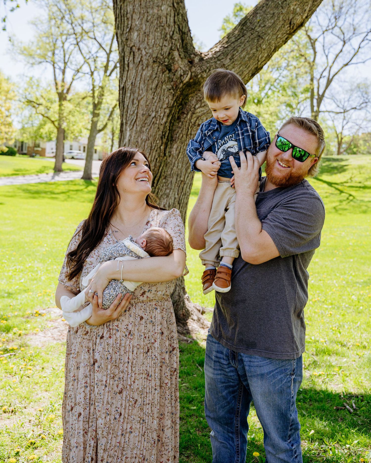 A couple shares a tender moment with their small dog while standing near a large tree trunk in a sunny park setting.
