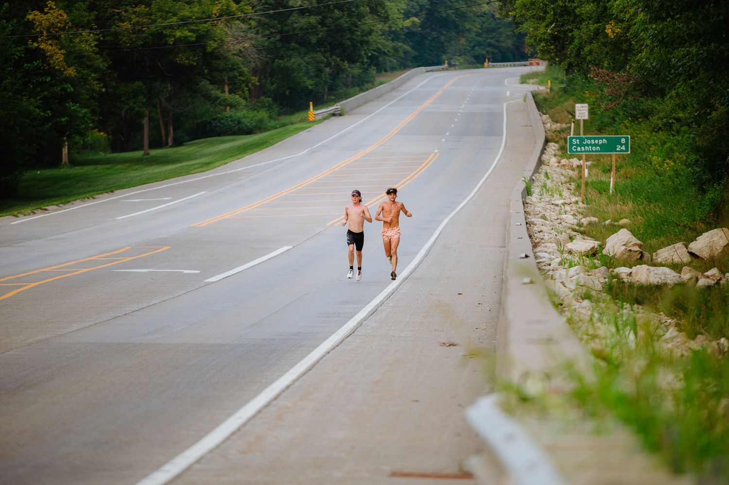Joggers running along a scenic rural road surrounded by green trees and nature.