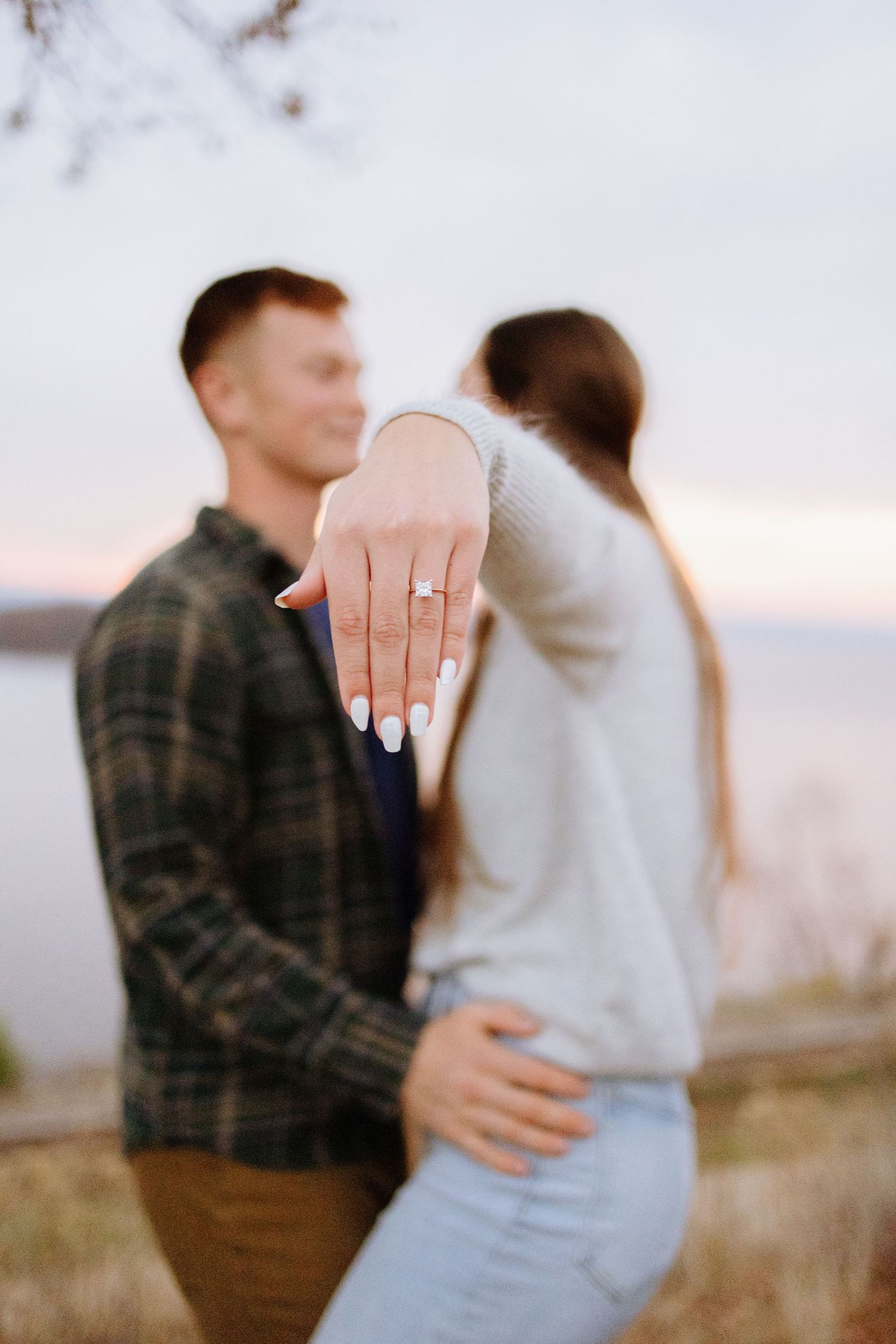 Close up of engagement ring with couple embracing in blurred background at sunset.