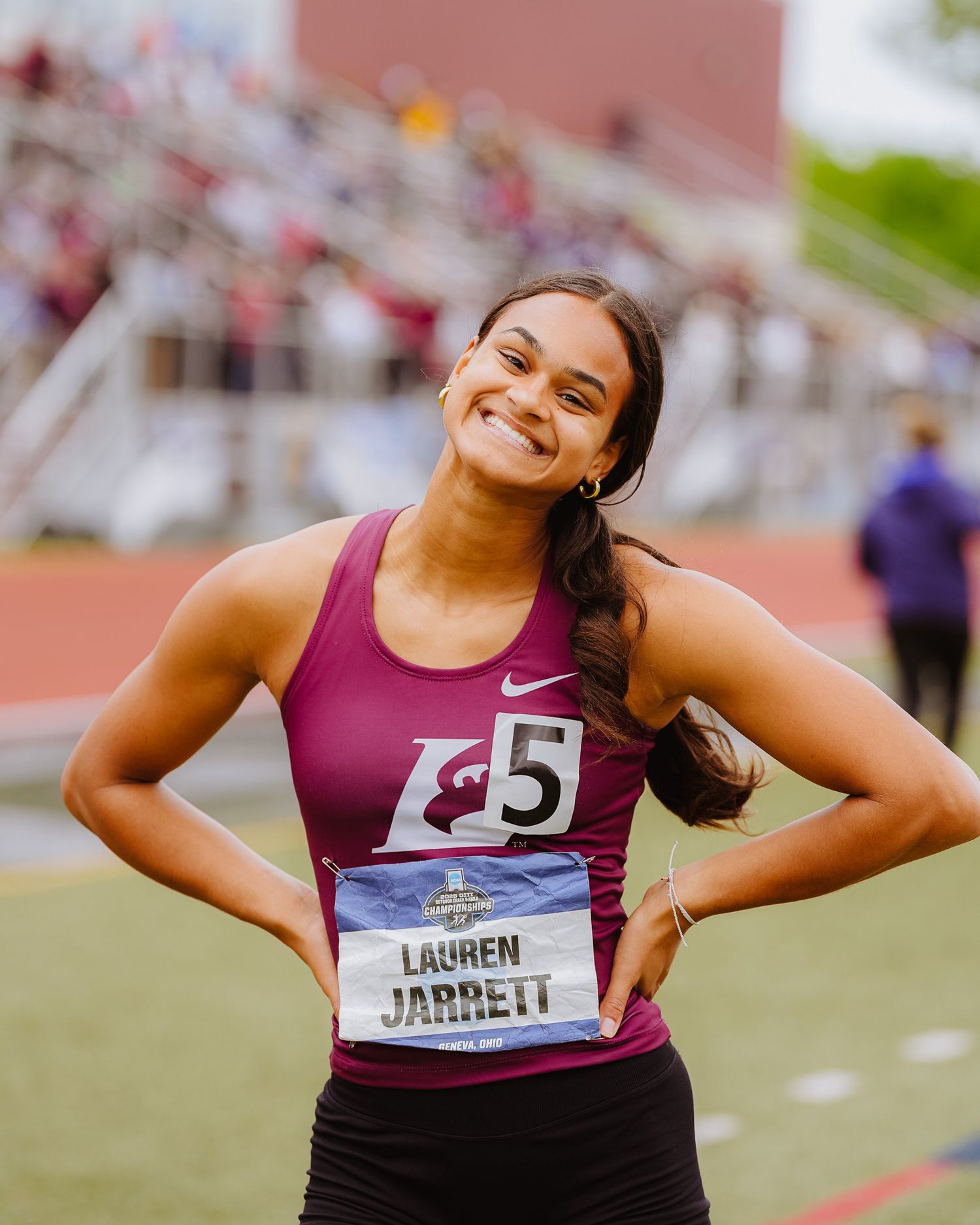 Athlete in purple tank top smiles joyfully on track field with stadium bleachers in background.