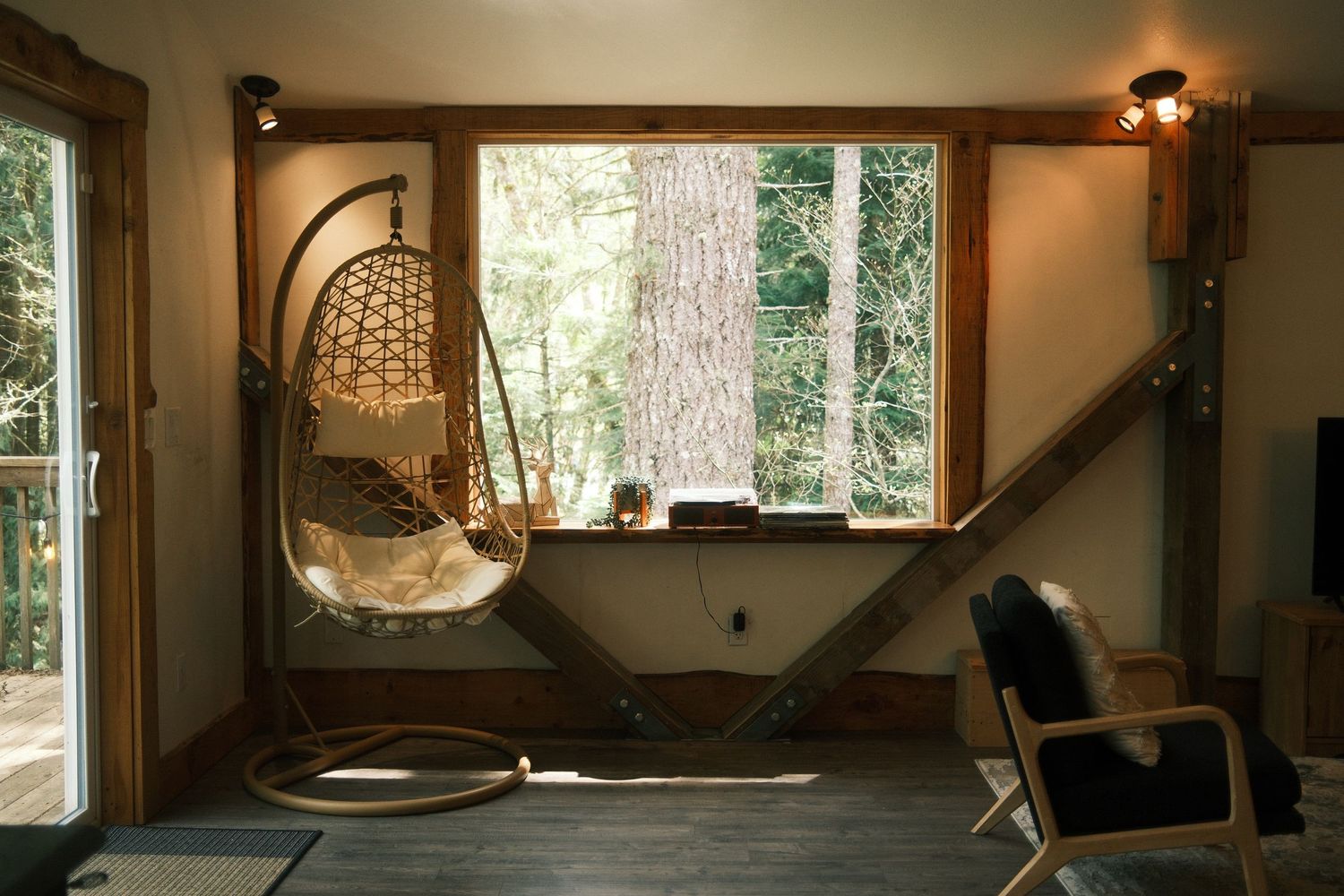 Cozy treehouse interior with hanging rattan egg chair, large window overlooking forest, wooden staircase, and warm lighting.